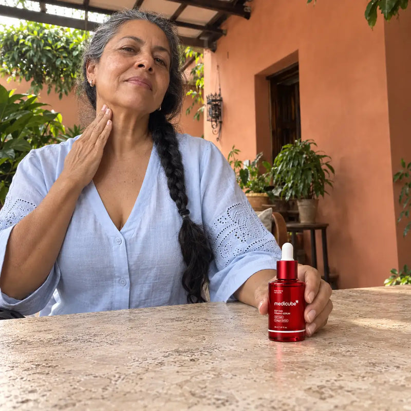 Mujer de trenza entrecana cuidando su cuello mientras sujeta el sérum apoyado sobre una mesa de piedra.